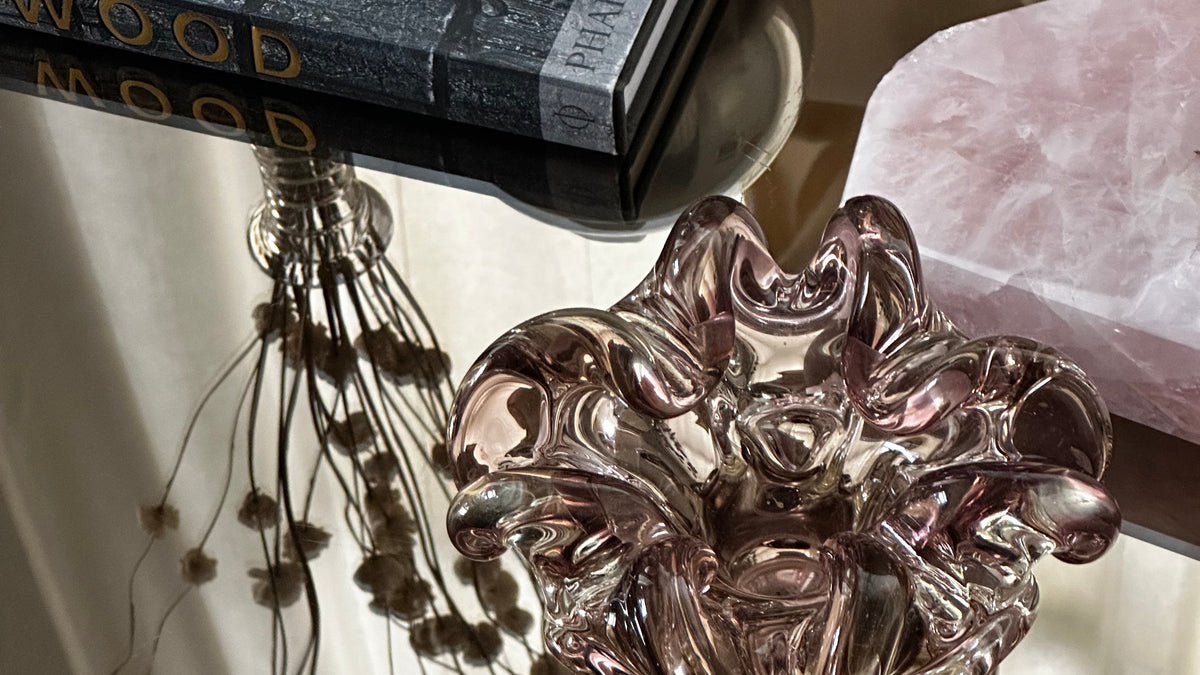 A decorative sculptural bowl made of iridescent, wavy translucent material in a pinkish-purple tone, displayed on a reflective surface next to a pink crystal and a dark textured book.